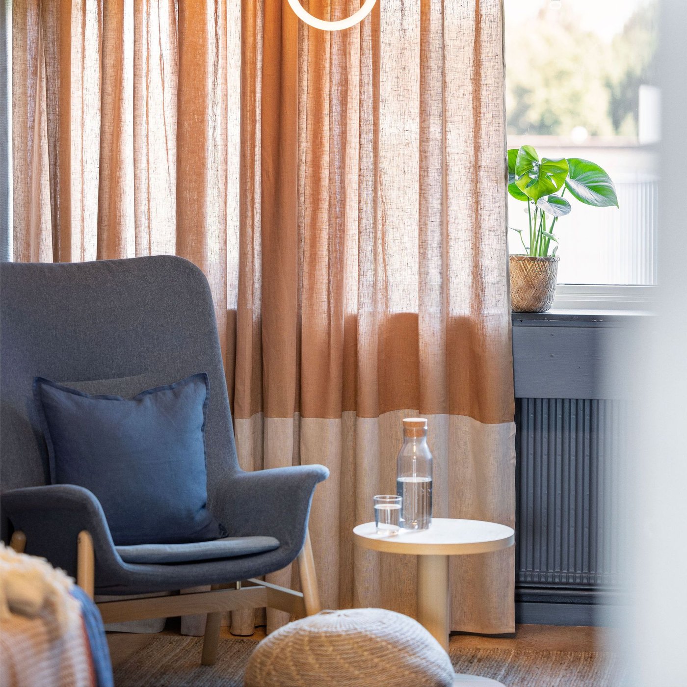 Mysig hörna med mörkgrå fåtölj och blå kudde, ett litet runt bord med vattenflaska och glas, samt beige gardiner och en grön växt i fönstret som släpper in dagsljus.