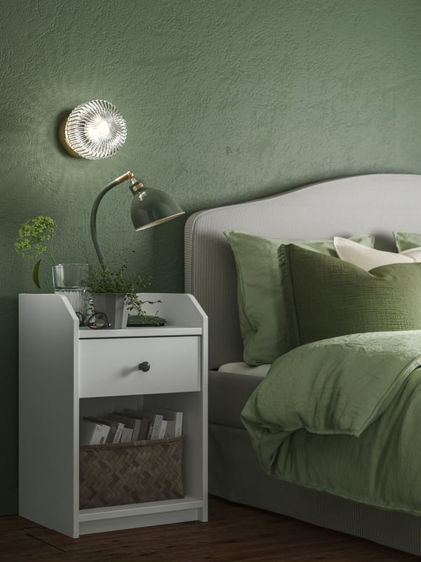 Close-up of HAUGA bedside table next to RAMNEFJÄLL upholstered bed with cushions and a duvet. All in green, grey and white.