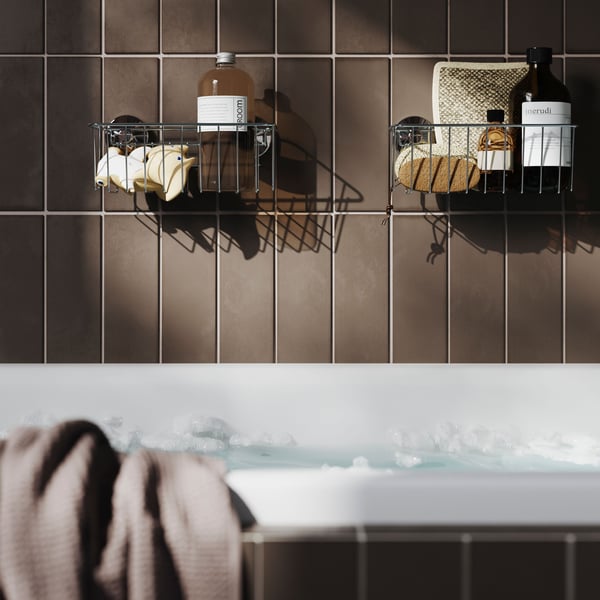 Zwei verzinkte KROKFJORDEN Körbe mit Saugnäpfen sind mit Toilettenartikeln gefüllt und stehen auf einer braun gefliesten Wand über einer Badewanne.