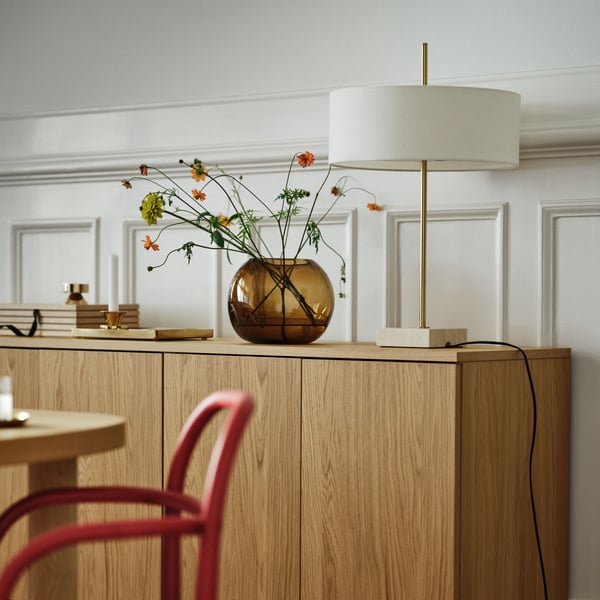 A brass-plated and white STOCKHOLM 2025 table lamp sits next to a brown vase on a cabinet by a white wall with wooden panels.