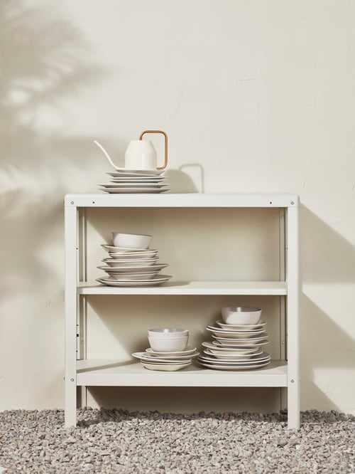 Une étagère d’intérieur/extérieur en acier laqué beige KOLBJÖRN se trouve sur du gravier, contre un mur. Un arrosoir et de la vaisselle sont posés dessus.