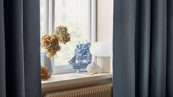 På fönsterkarmen står en DEJSA bordslampa, en liten vit vas och en större vit vas med blommor. Gardinerna är gråa och hänger vid fönstret.