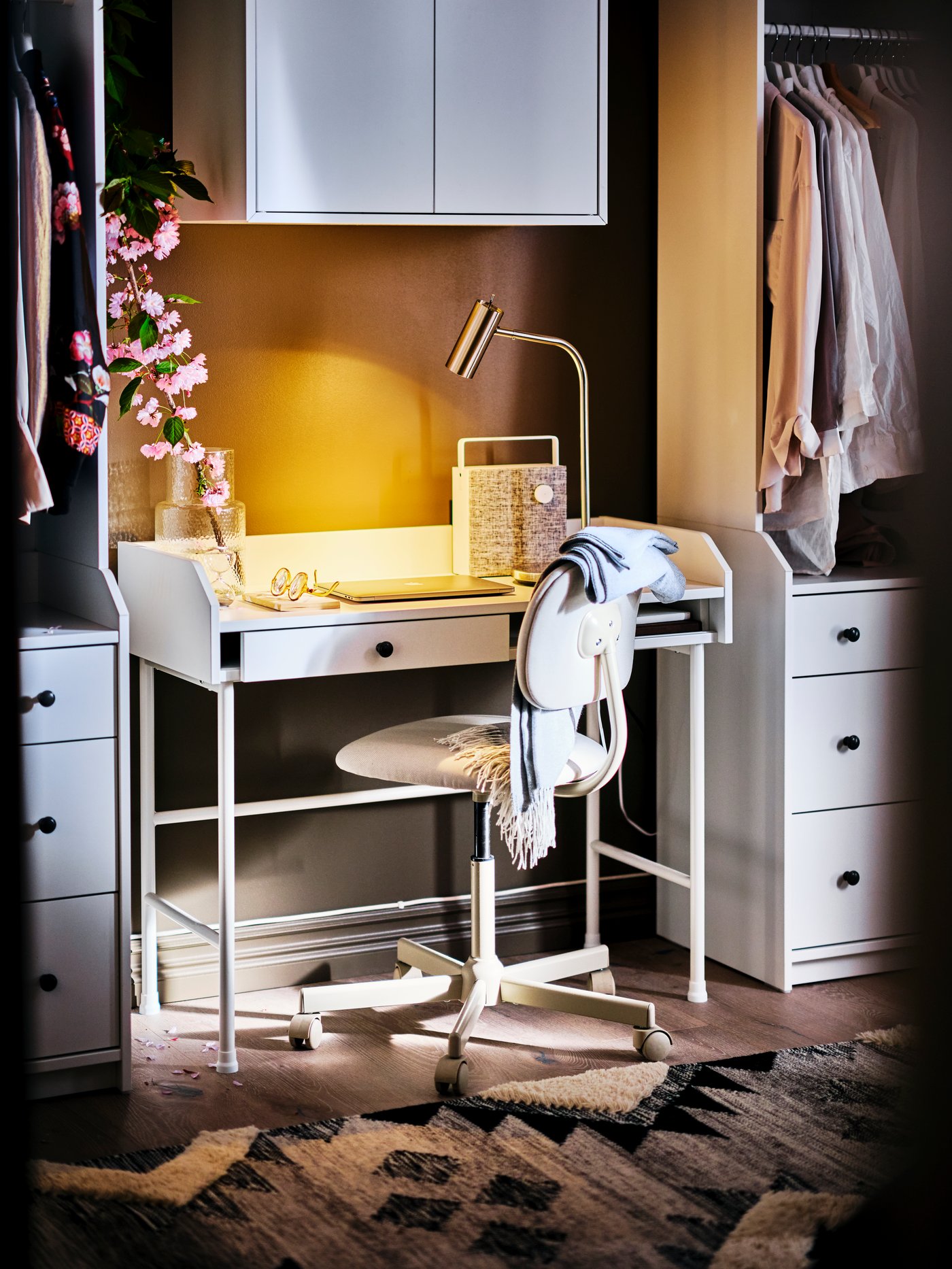 Espacio de trabajo con un escritorio rectangular, un mueble con dos puertas, una silla giratoria y una cómoda con un estante y ropa.