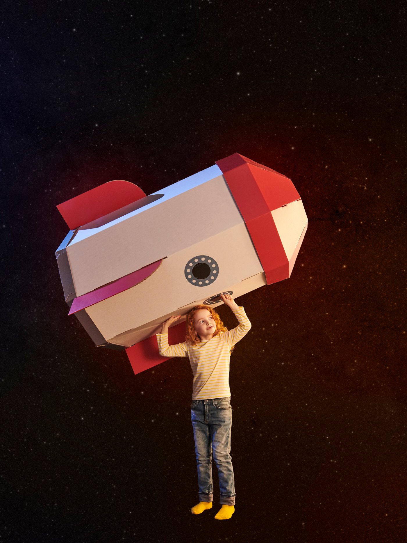 A child in a galaxy-themed space holds an AFTONSPARV rocket children’s tent on its side over her head, pointing its nose up.