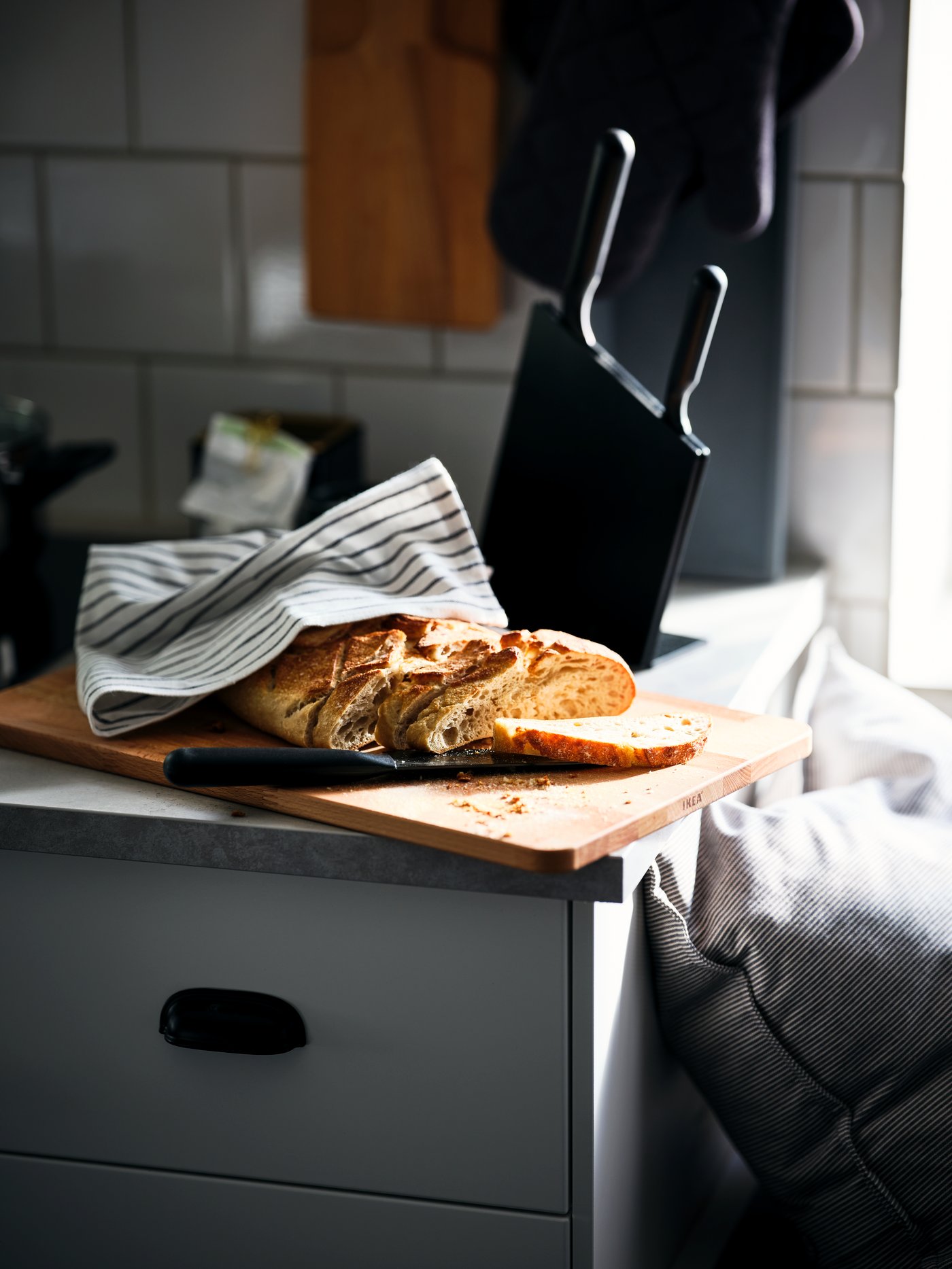 A patterned RINNIG tea towel enveloping a fresh loaf of bread by a black JÄMFÖRA three-knife block on a sunlit worktop.