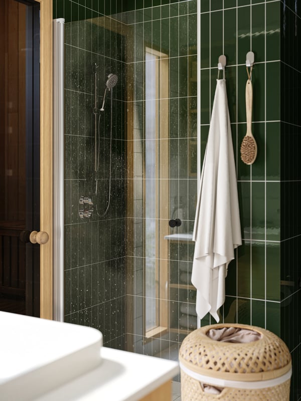 A shower is placed in a green-tiled bathroom and there is a white FREDIKSJÖN towel hanging from hooks on the wall next to it.