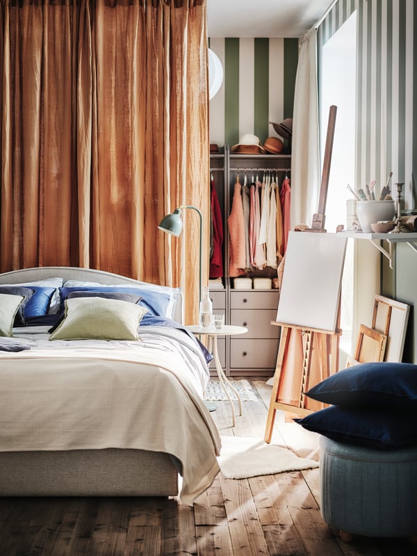 A beige RAMNEFJÄLL bed with dark beige DYTÅG curtains and a lamp behind it and various cushions on top, some in DYTÅG covers.