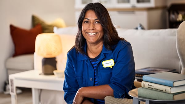 Une designer d'intérieur IKEA dans sa tenue de travail bleue est assise dans une pièce d'exposition et sourit à la caméra.