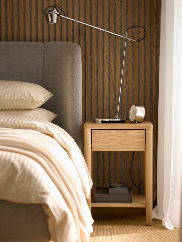An oak veneer TONSTAD bedside table with a chrome-plated MODERMOLN work lamp on top beside a TUFJORD upholstered bed.