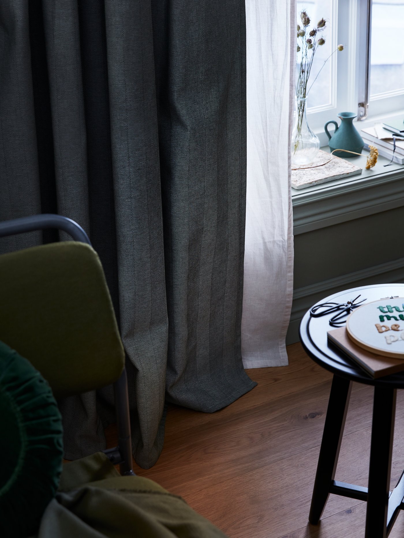 Des rideaux assombrissants TIBAST vert foncé sont installés par-dessus des rideaux HANNALILL plus légers à une fenêtre. Des livres décorent le rebord de la fenêtre.