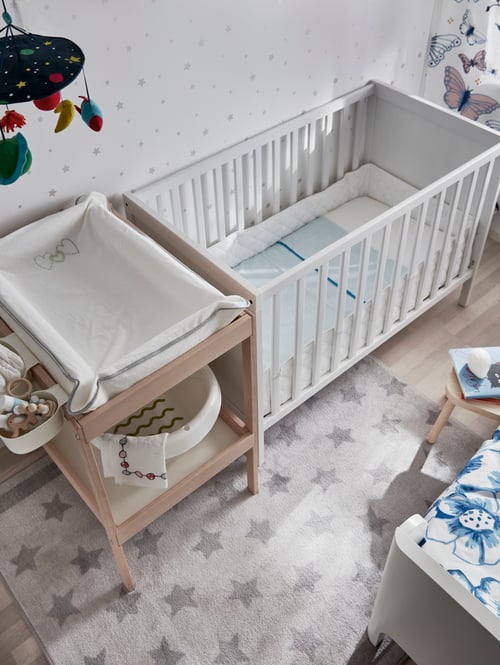 Modern nursery with white crib next to changing table, rug, children's stool, baby care mat and toys.