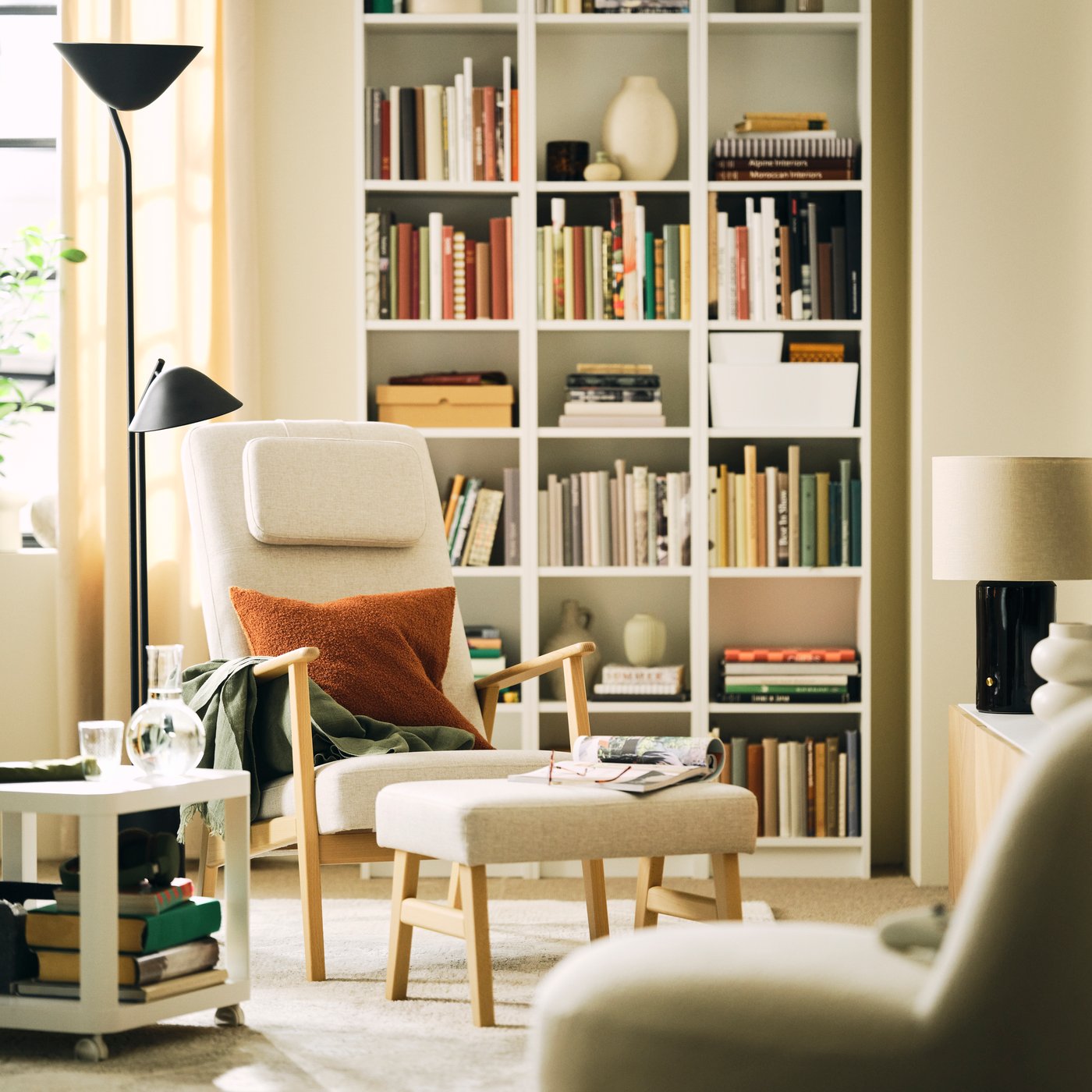 A living room with a light-beige EKENÄSET high-back armchair and footstool beside a white bookcase and a lamp.