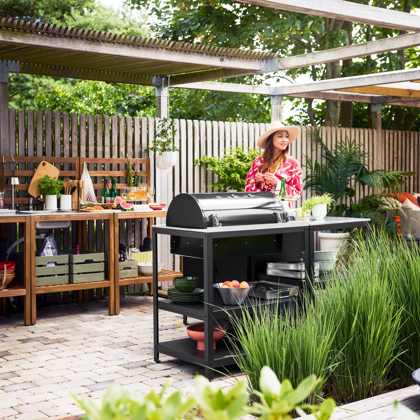A patio with greenery showcases a black GRILLSKÄR charcoal barbecue on a kitchen island, a woman standing next to it.