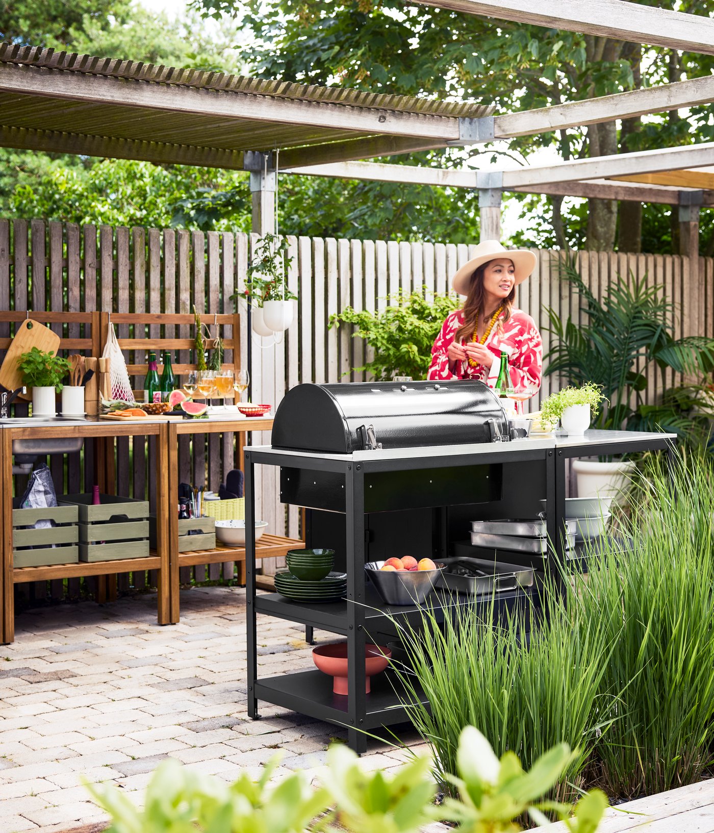 En terrasse med grønne områder fremviser en sort GRILLSKÄR kulgrill på en køkkenø, med en kvinde der står ved siden af.