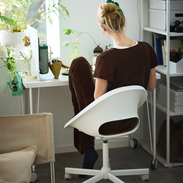 A person dressed in brown sitting on a white LOBERGET swivel chair by a white desk with green plants by a window.