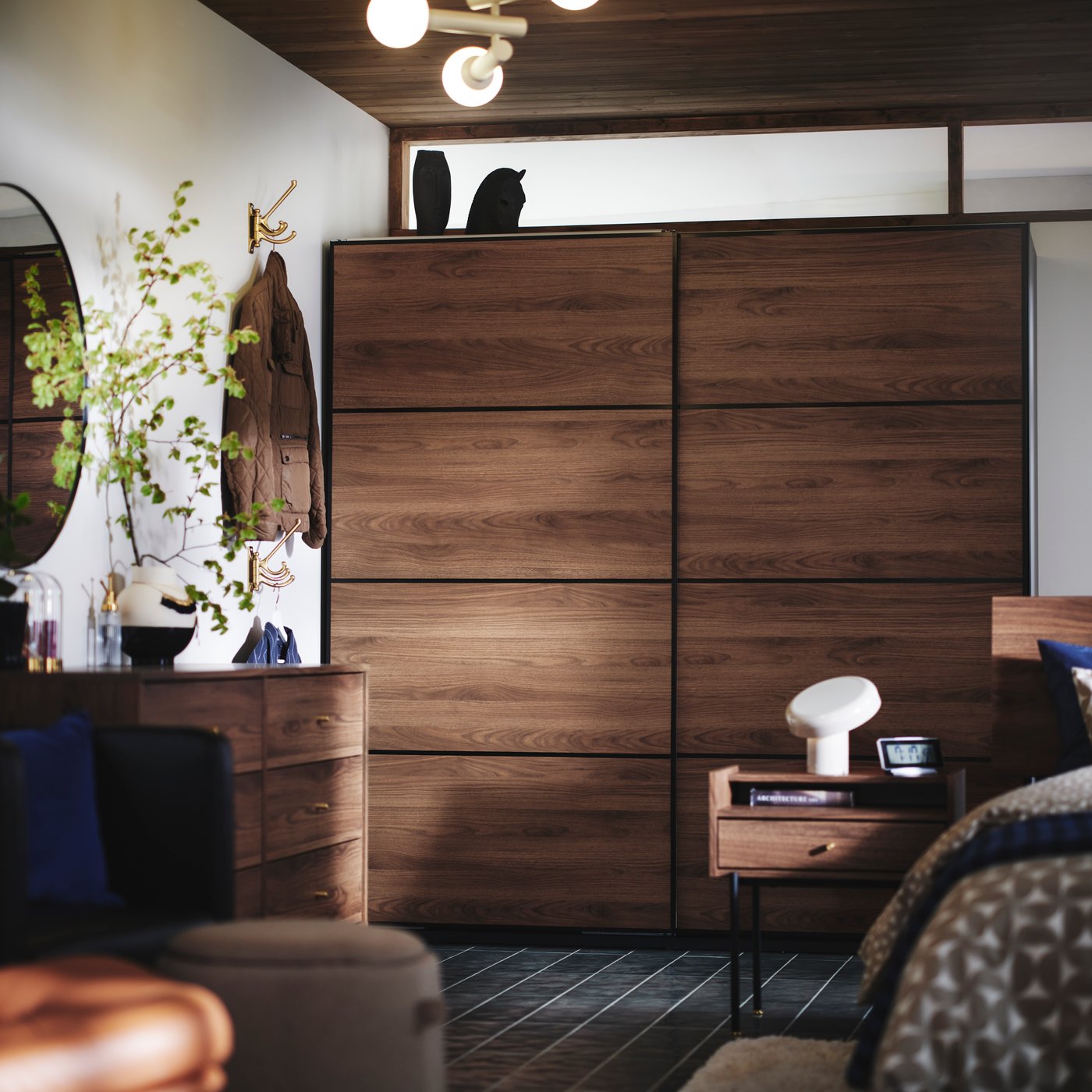 The side of a bed, RÅDMANSÖ bedside table and PAX MEHAMN wardrobe in dark wood and sliding doors in the background.