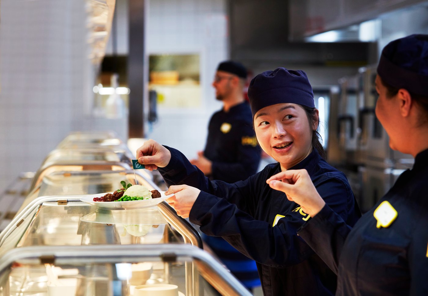 Tummansinisiin työasuihin pukeutuneet IKEA-työntekijät annostelevat aterioita ravintolassa.