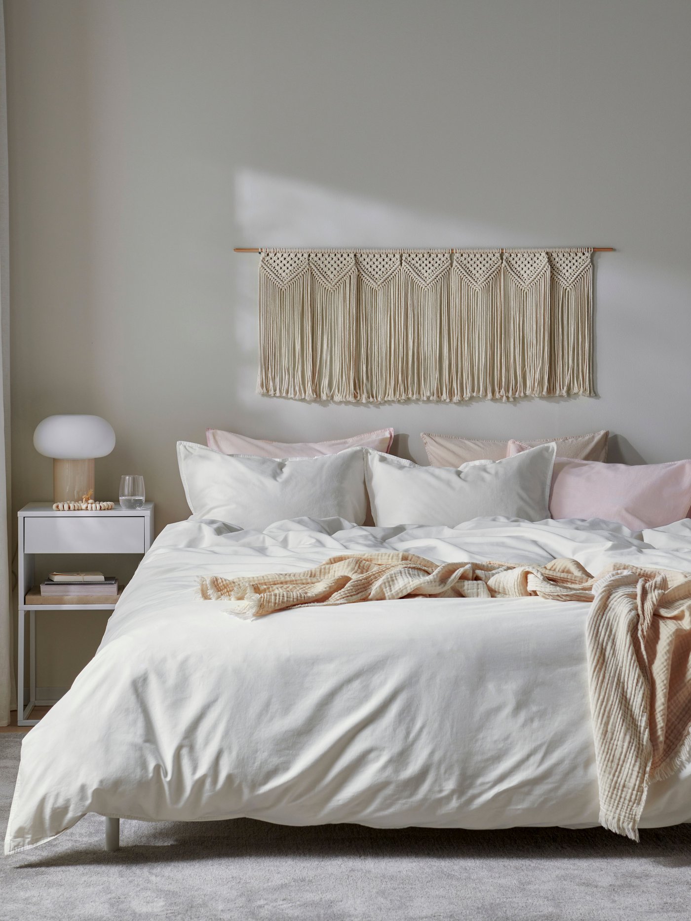 Una camera da letto luminosa dai toni chiari con un copripiumino e federa ÄNGSLILJA bianchi. Sopra il letto, ci sono cuscini e plaid bianchi e beige.