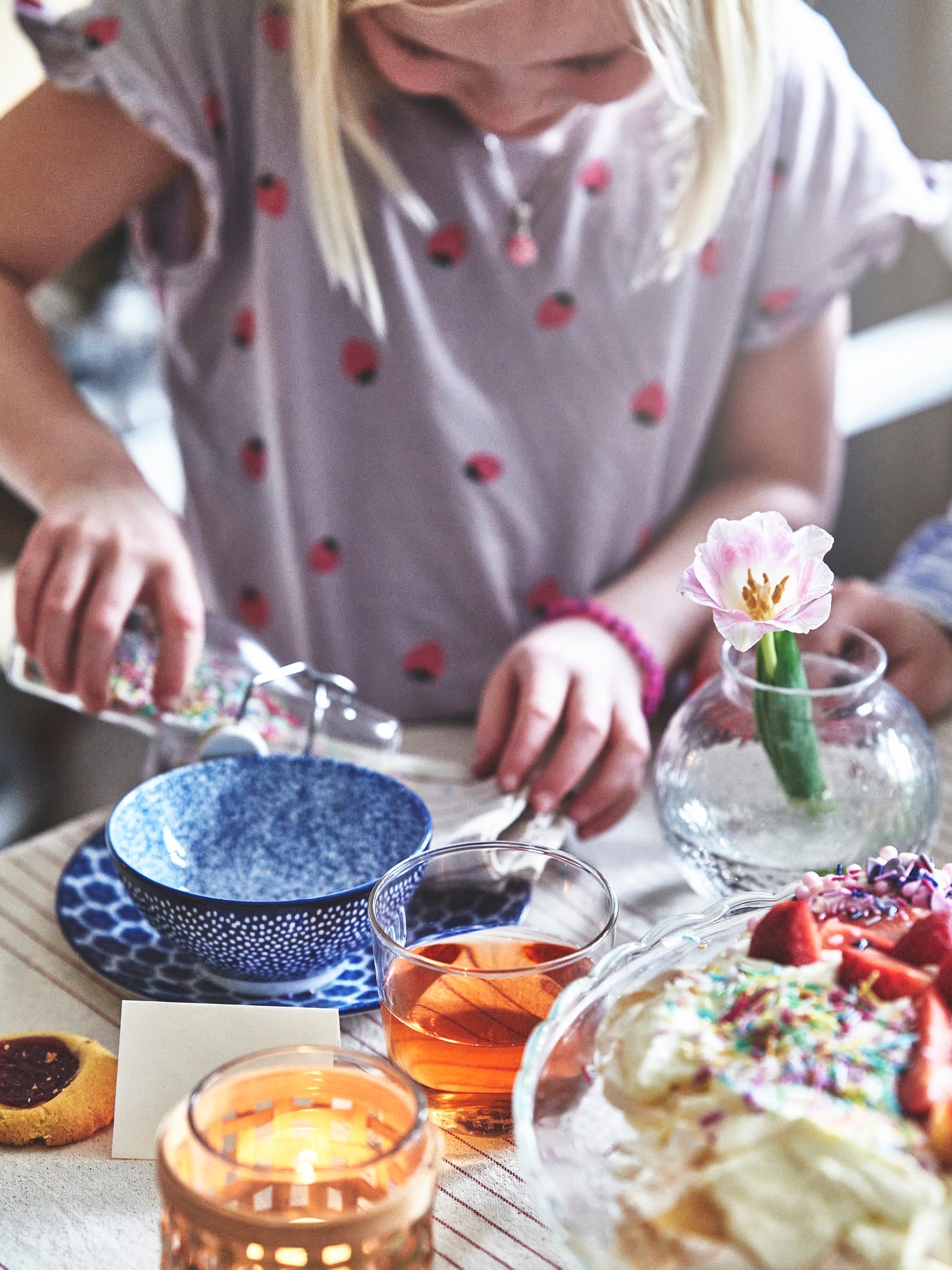 Närbild av ett dukat bord med desserttillbehör, där ett barn häller något i en blåvit skål. På bordet finns glas med dryck, kakor, jordgubbar, vispad grädde och ett litet glas med en blomma.