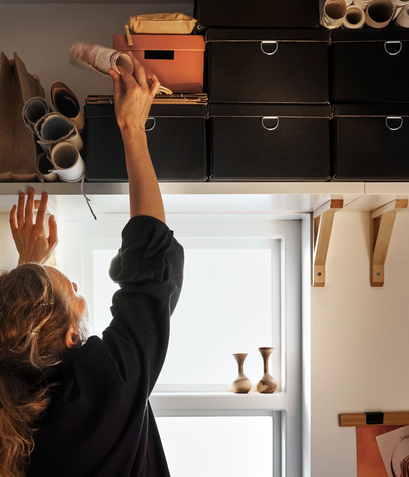 A person reaches for an item beside stacked boxes on a white BERGSHULT wall shelf with aspen brackets mounted over a window.