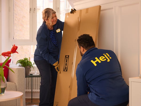 Deux livreurs transportent un colis IKEA en kit dans un salon lumineux où ils assurent également le montage des meubles.