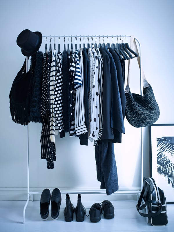 A bedroom with bluish tints. A MULIG clothes rack is standing with clothes in dark tones of black and blue hangin on it. There are 3 pairs of shoes on the floor next to a frame against the wall and a bag.