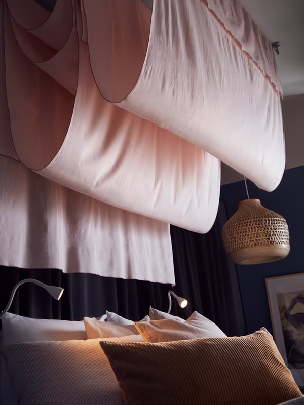 Pink MOALISA curtains hanging in waves from the ceiling next to a bamboo pendant, above a bed with grey striped bed linen.