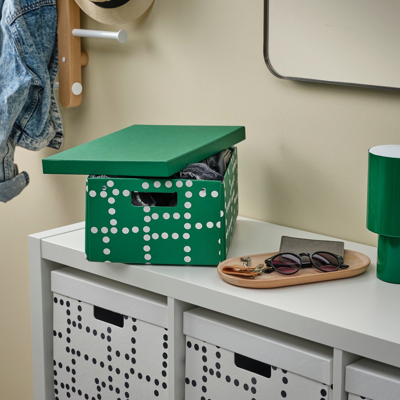 A green lidded storage box with a white dotted pattern sits open on a white entryway cabinet, partially filled with clothing. Matching white storage boxes with the same dotted pattern are stored in the cubbies below, with a tray holding keys, sunglasses, and a wallet, plus a green table lamp nearby.
