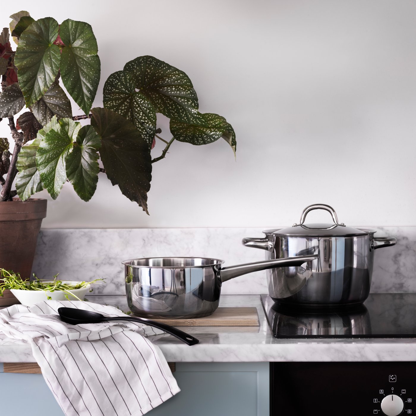 A stainless-steel saucepan and pot sit on a marble countertop beside a potted plant, towel, and spoon in a modern kitchen.