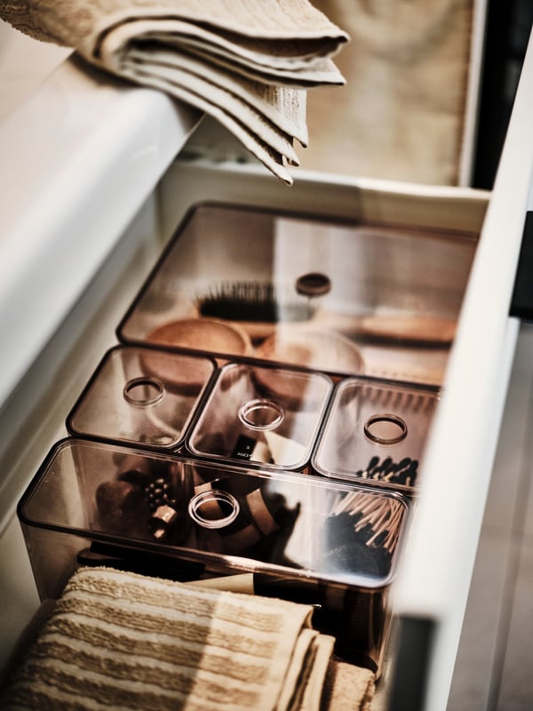 An open washstand drawer revealing a light beige VÅGSJÖN bath towel and smoked GODMORGON boxes with lids holding accessories.