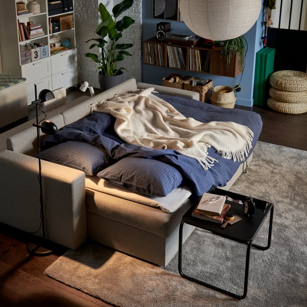 Dans un salon plongé dans une demi-obscurité, un canapé convertible BÅRSLÖV doté d’une housse de couette et de taies d’oreiller DOFTAKLEJA est prêt pour la nuit.