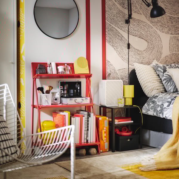 A bright red KRÖNIKÖR open storage unit holds various items against a stylish patterned wall by an upholstered bed.
