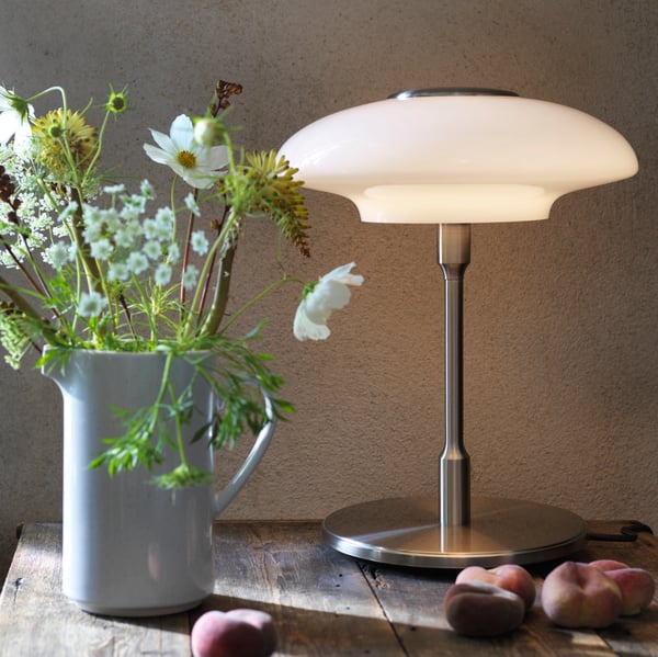 A table lamp on a wooden table, beside a white colored jug filled with plant stems and flowers.