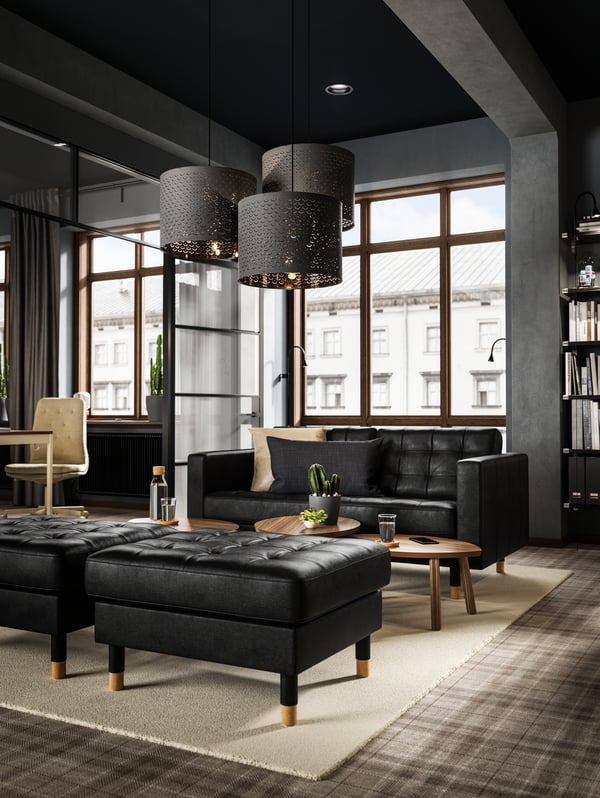 In een ruim appartement met een donker industrieel interieur staan er twee voetenbanken in zwart leer vóór een LANDSKRONA zitbank in zwart leer.