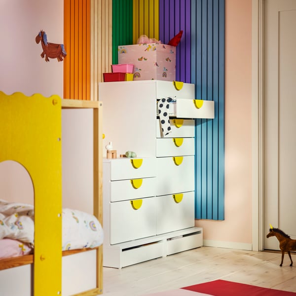 A SMÅSTAD/PLATSA chest in white with yellow handles and open shelves, in a colourful children’s room with striped walls.