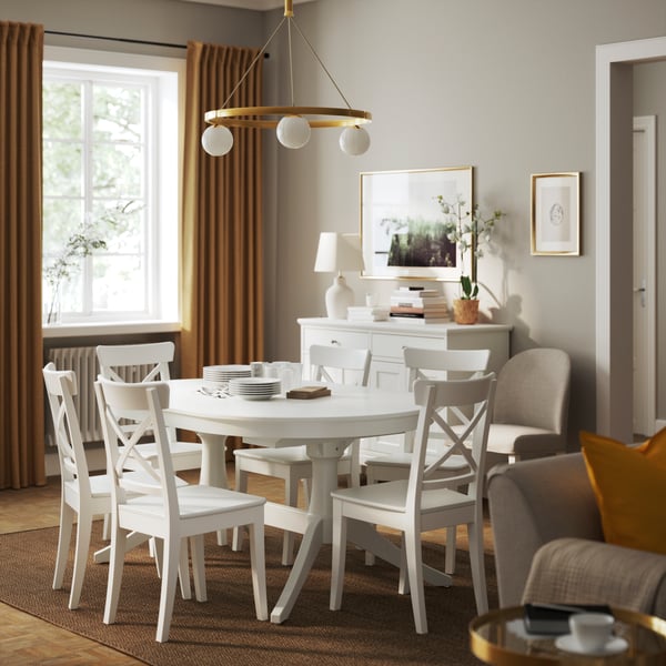 Gemütliches Esszimmer mit einem runden weißen ROSENTORP Tisch und sechs weißen ROSENTORP Stühlen sowie einem goldenen Kronleuchter. Warmes Sonnenlicht fällt durch ein Fenster mit senffarbenen Vorhängen. Neutrale Einrichtung.