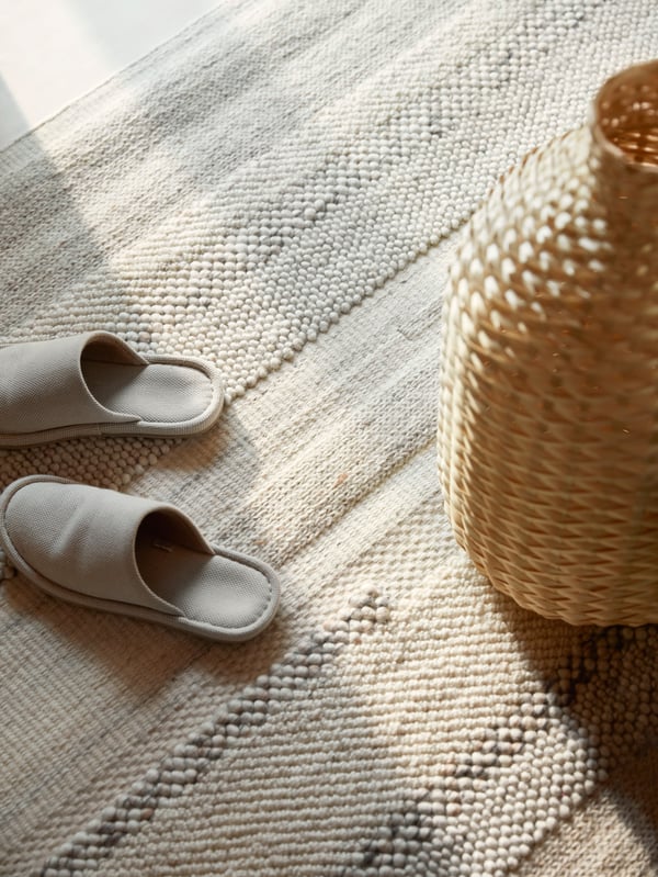 Cream-coloured slippers and a woven bamboo vase stand on a hand-woven BRÖNDEN rug.