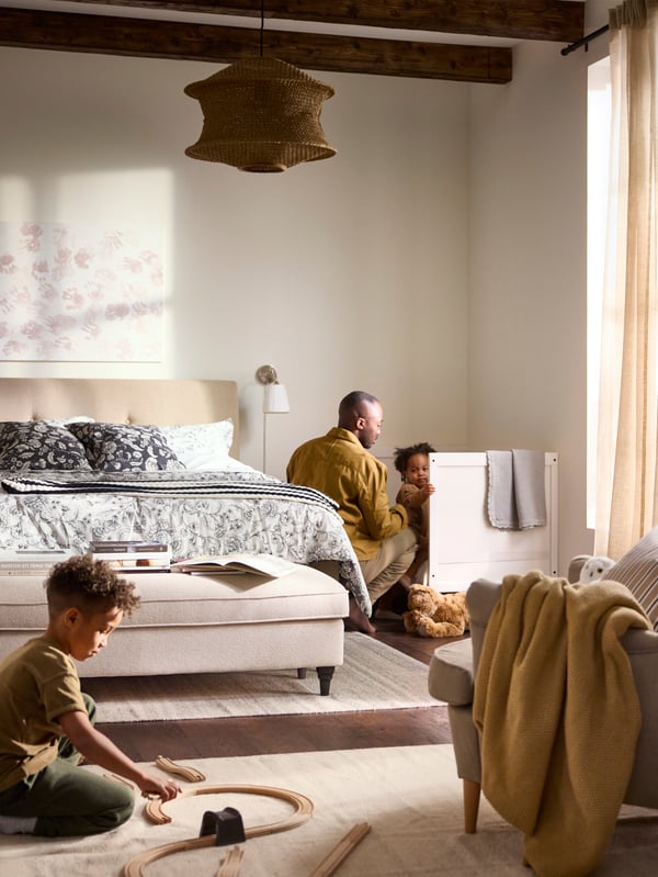 Bedroom in beige and white, a child playing with a train set on the floor, another child being put to bed by parent in back.