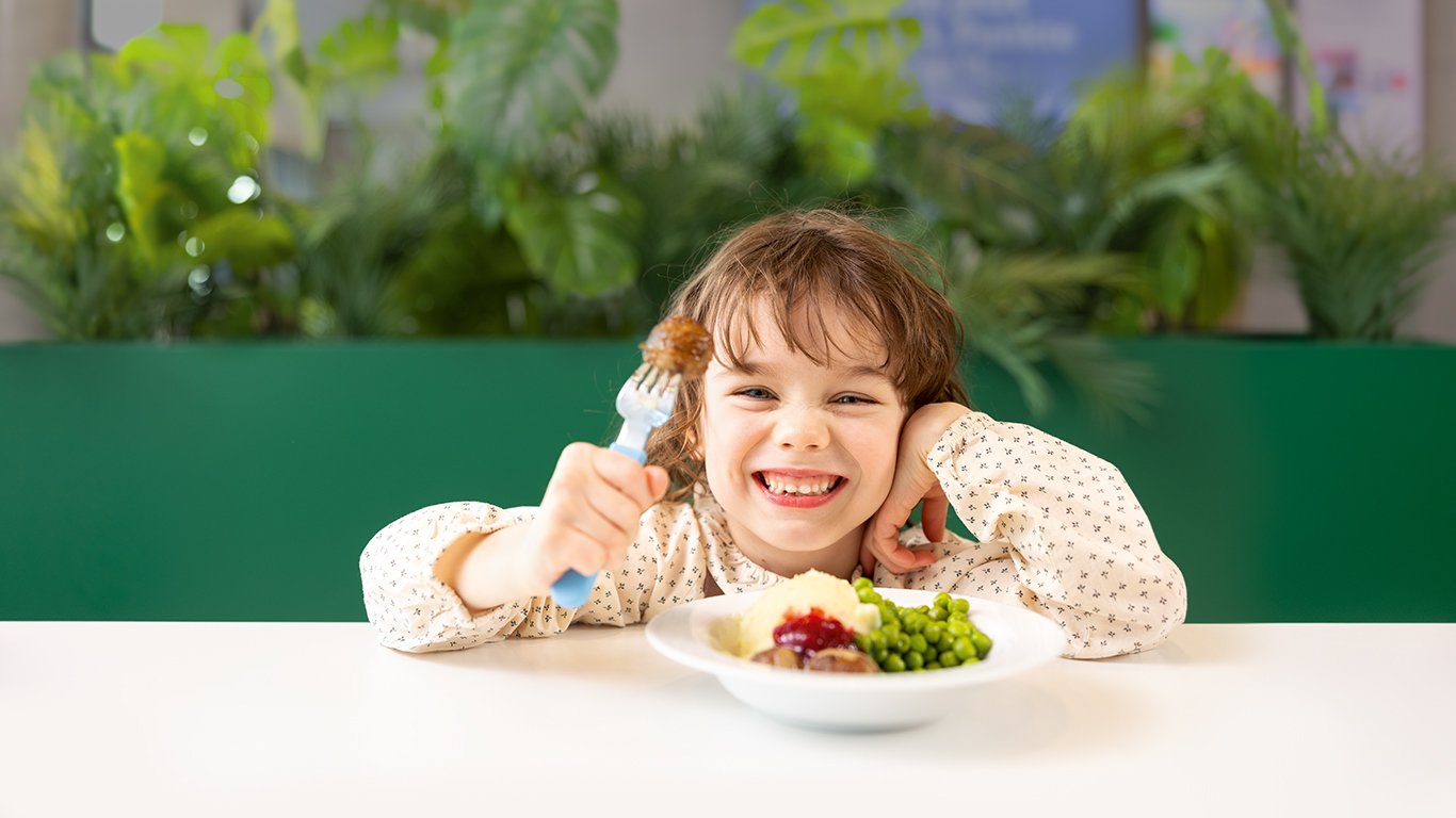 Ein Kind sitzt an einem Tisch im IKEA Restaurant vor einem Teller mit Fleischbällchen und hält eine Gabel in die Kamera.  Im Hintergrund sorgen grüne Pflanzen und helle Farben für eine freundliche Atmosphäre.