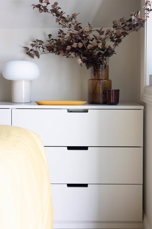NORDLI kommode med en lampe, en vase med blomster og et fat på toppen.
