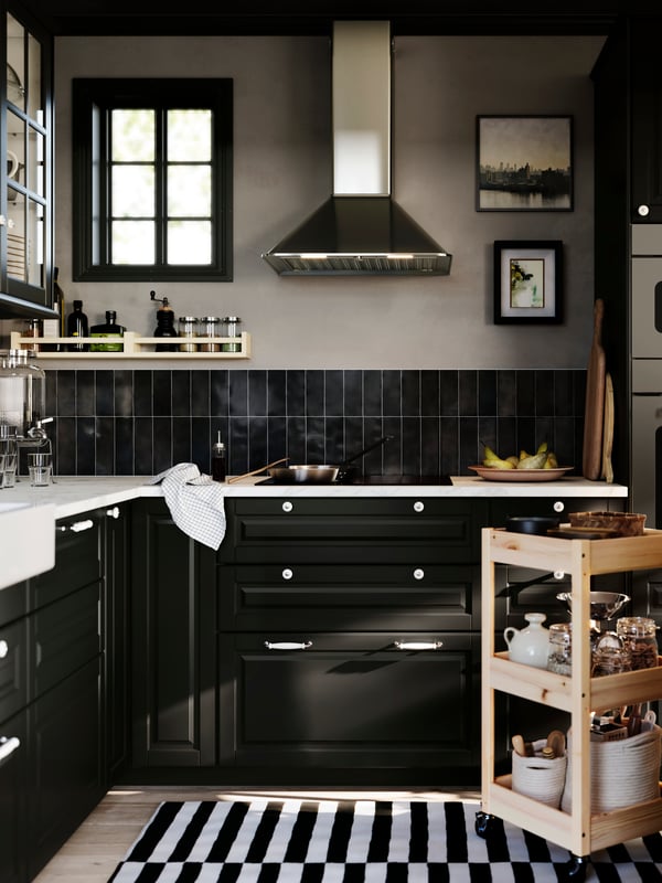 An L-shaped kitchen with black BODBYN drawers has a hob and a MATTRADITION wall mounted extractor hood in stainless steel.
