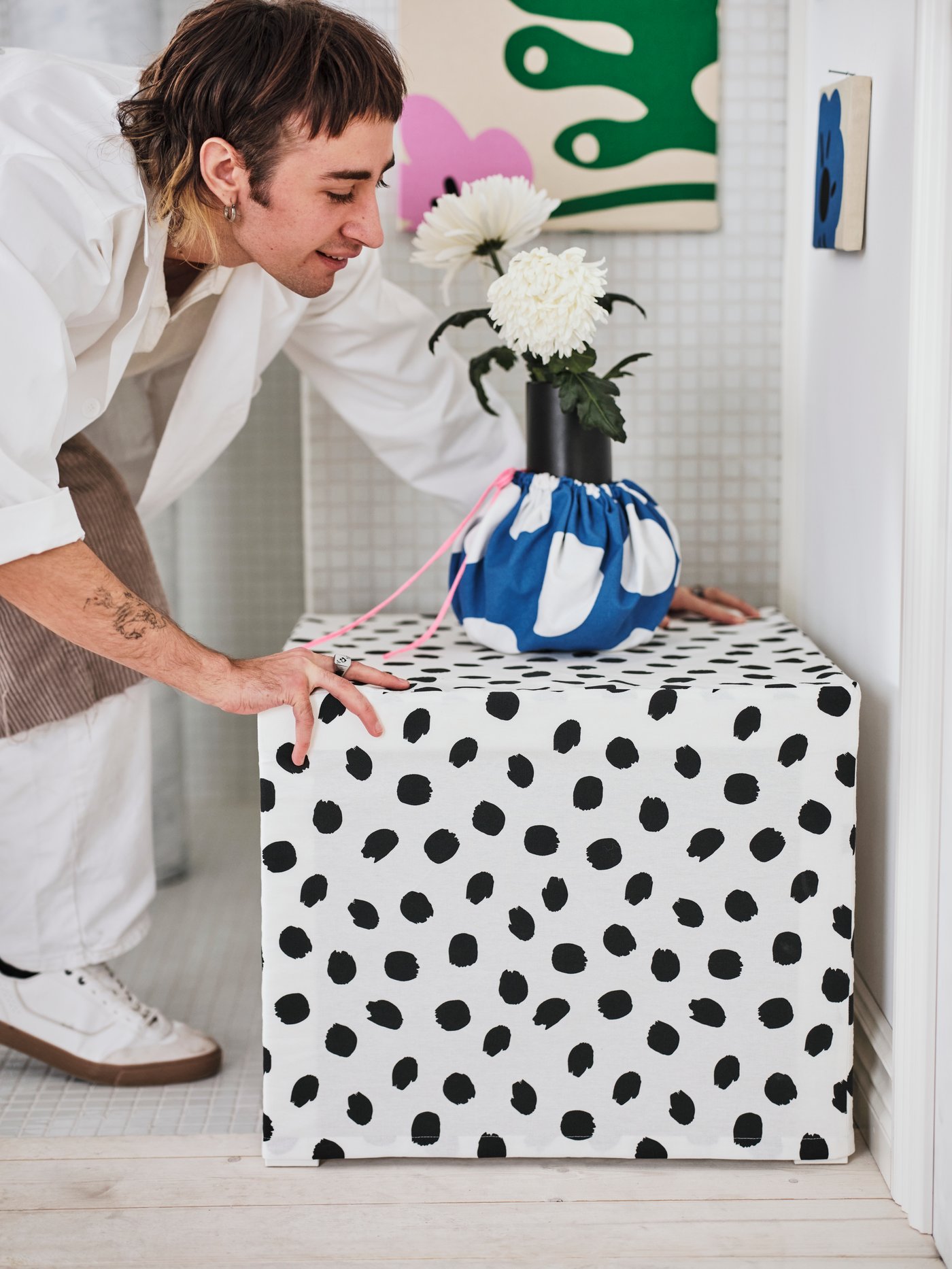 A person adjusts a small side table covered with the SANDLUSERN pre-cut fabric. On top of the table is a fabric-covered vase.