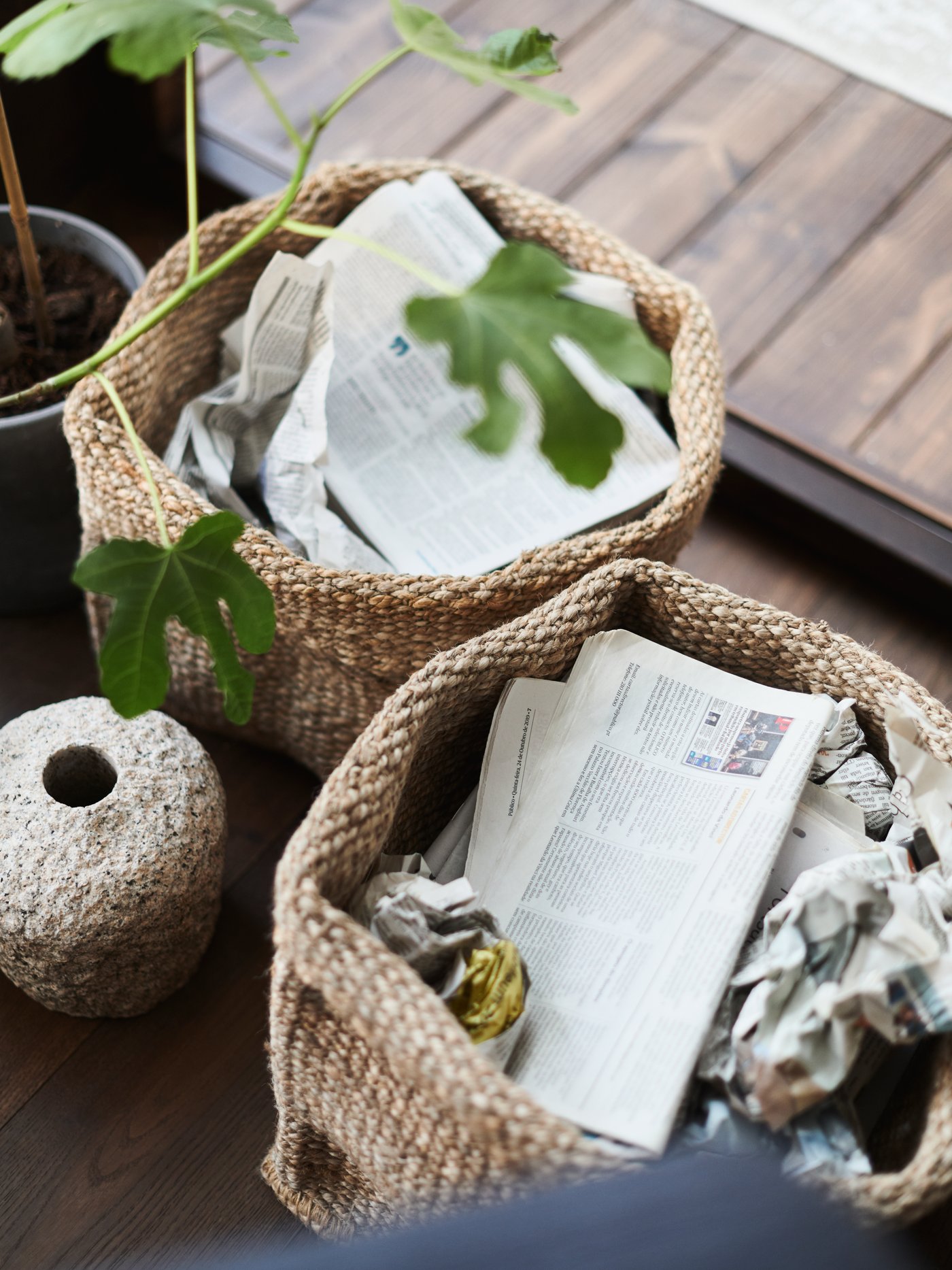 Two TJILLEVIPS baskets on a wooden floor filled with paper, beside a small vase and a plant.