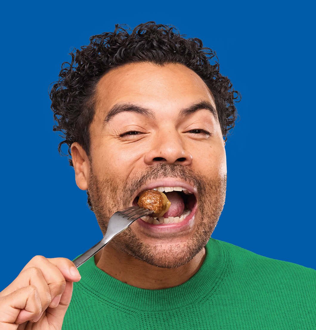Homme heureux avec un pull vert sur fond bleu, qui mange une boulette Suédoise avec une fourchette