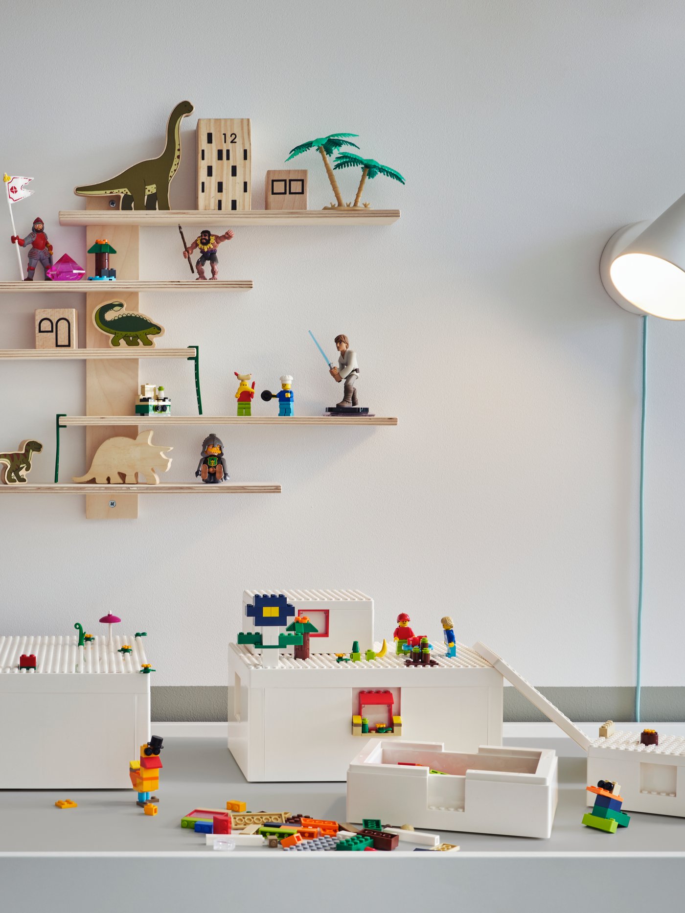 Una repisa de madera LUSTIGT instalada en un muro blanco sobre un escritorio cubierto por piezas de legos BYGGLEK. Sobre la repisa hay figuras de madera en forma de edificios y dinosauros. 