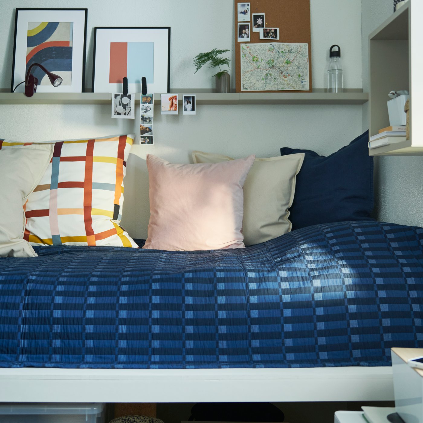 A dorm room with a blue SKOGSKOVALL bedspread laid over a bed, alongside cushions of various colours and a shelf above.