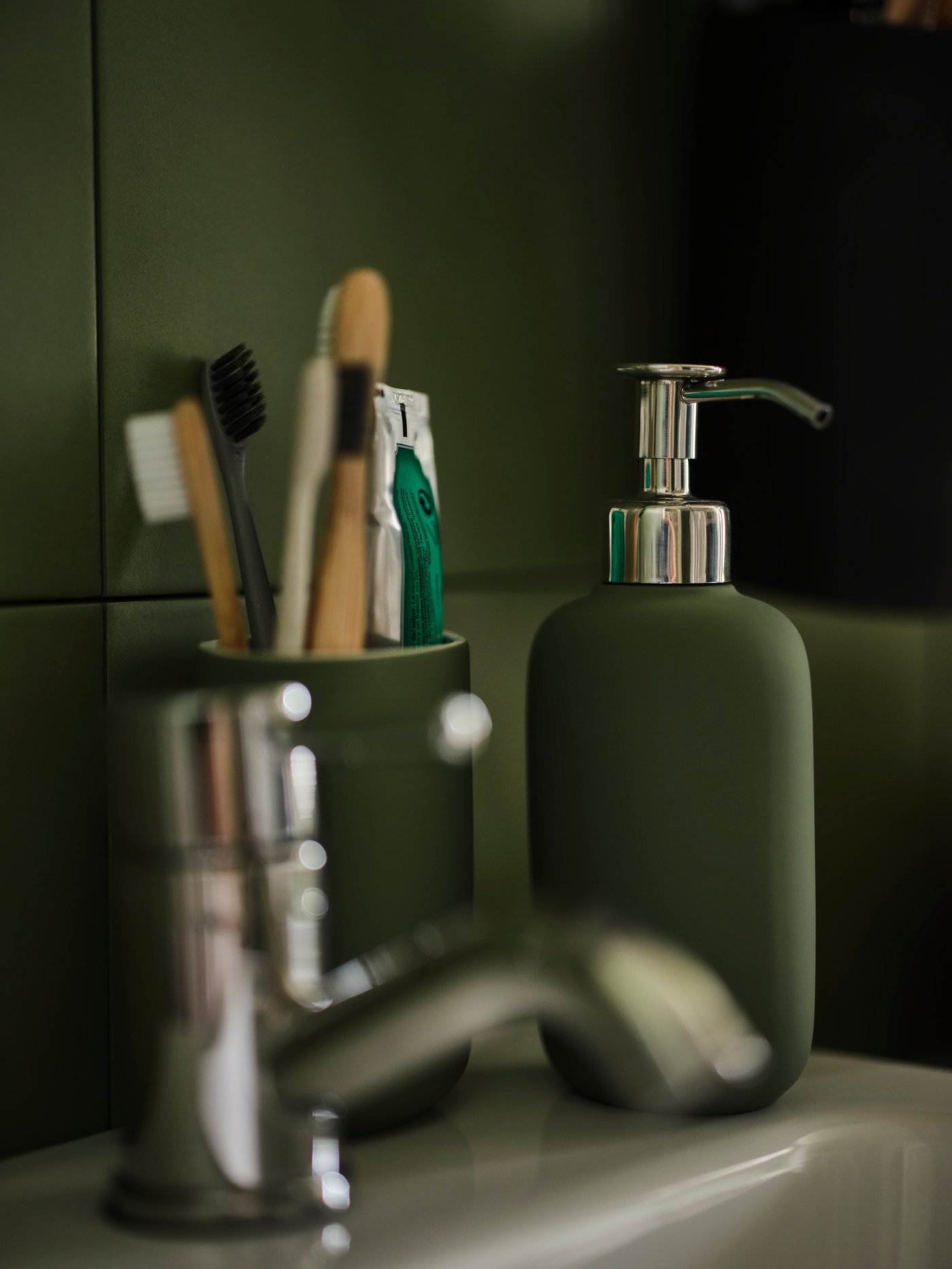 An EKOLN soap dispenser in grey-green, on a sink with a tap and a grey-green EKOLN toothbrush holder with toothbrushes.