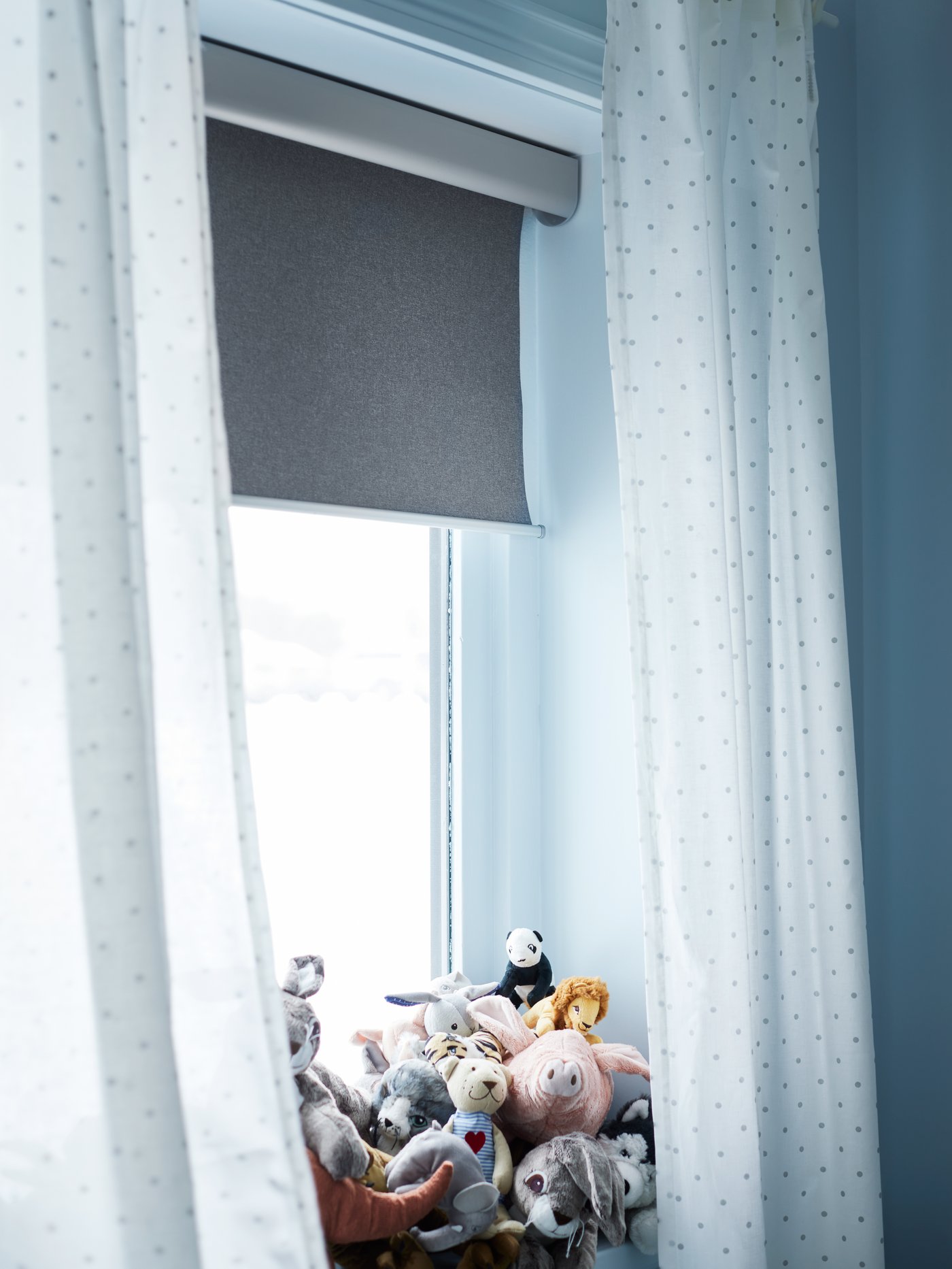Dormitorio infantil con una ventana grande con cortinas blancas, peluches en el alféizar y estor opaco gris claro sin cuerda.