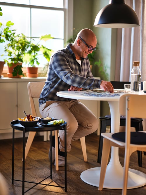 A man sitting on an ash LISABO chair at a white DOCKSTA table, building a puzzle with a black GLADOM tray table to his right.