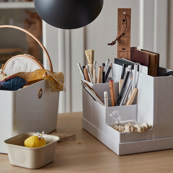 Una superficie con sopra tre contenitori di cui uno contentente materiali tessili, uno piccolo con dentro gomitoli con spilli e uno con materiali da disegno.